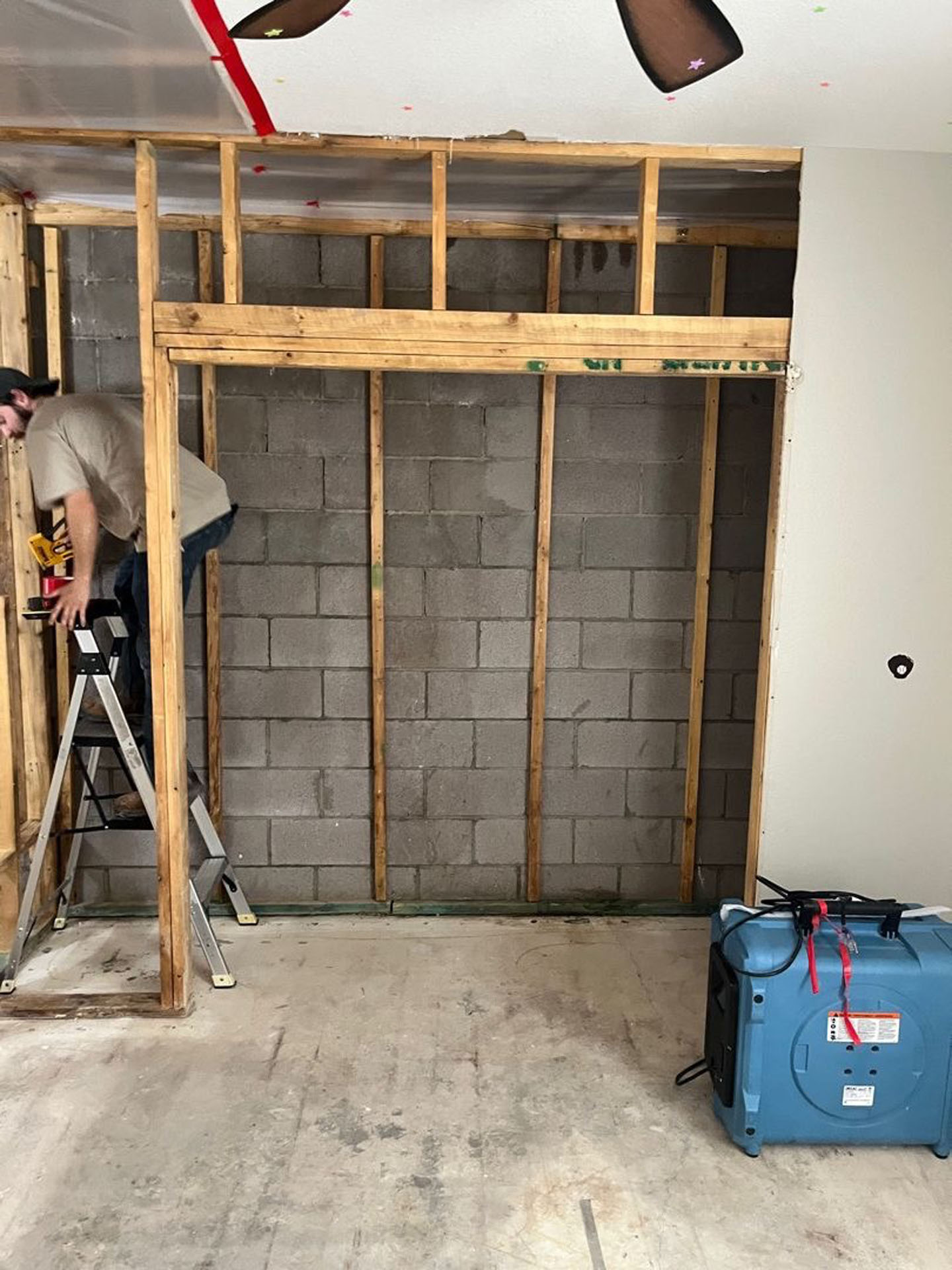 A contractor on a ladder installs wood framing against a concrete block wall during a renovation.