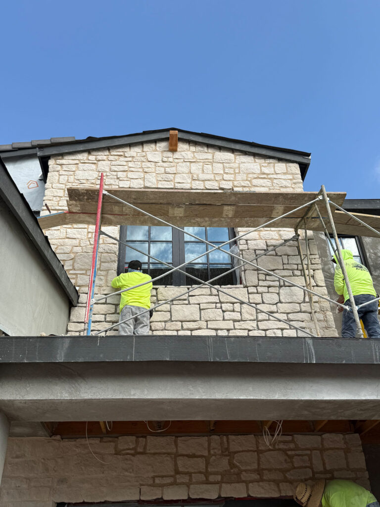 Construction workers on scaffolding installing light tan stone veneer on a residential house exterior.