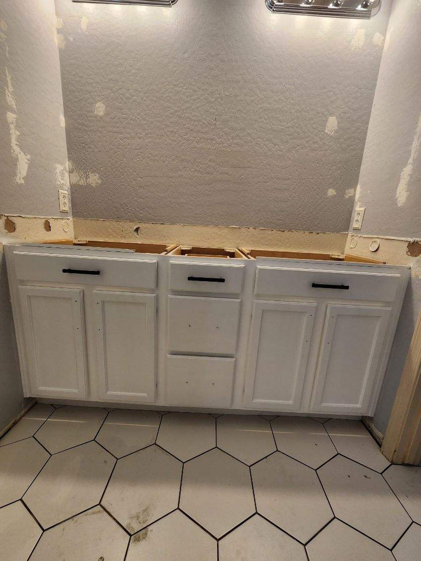 White bathroom vanity with black hardware installed over grey hexagon floor tiles during a renovation.