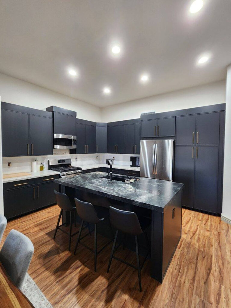 Matte black kitchen cabinets with gold hardware and a black marble island under recessed ceiling lights.
