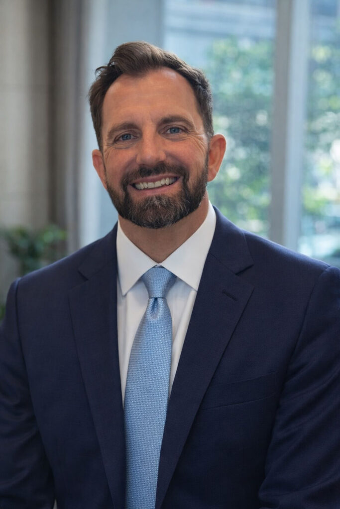 Professional man with a beard smiling in a navy blue suit and light blue tie in a bright office.