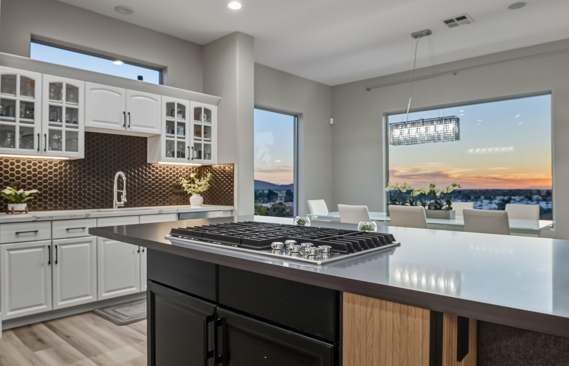 Modern kitchen featuring a grey island with gas cooktop, white cabinetry, and sunset views through large windows.