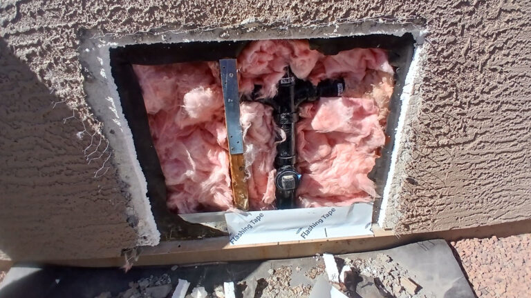 Exposed pink fiberglass insulation and black plumbing pipes inside an opening in a brown stucco wall.