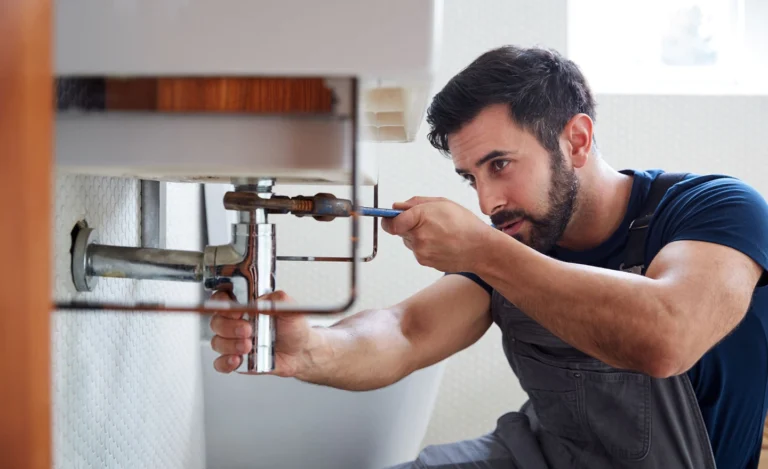 A professional plumber in grey overalls uses a wrench to repair a chrome sink pipe during a remodel.