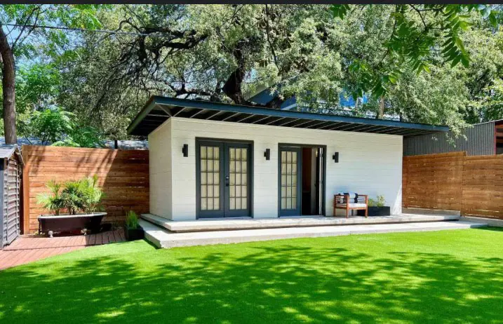 Modern white backyard studio with black trim, French doors, and a wooden fence on a green lawn.