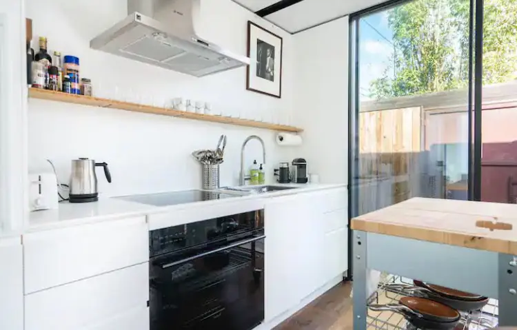 Modern white kitchen featuring minimalist cabinetry, a black oven, stainless steel hood, and a wooden floating shelf.