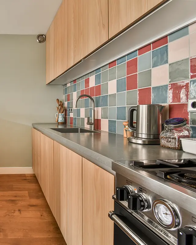Modern kitchen with light wood cabinets, colorful square tile backsplash, and stainless steel countertops.